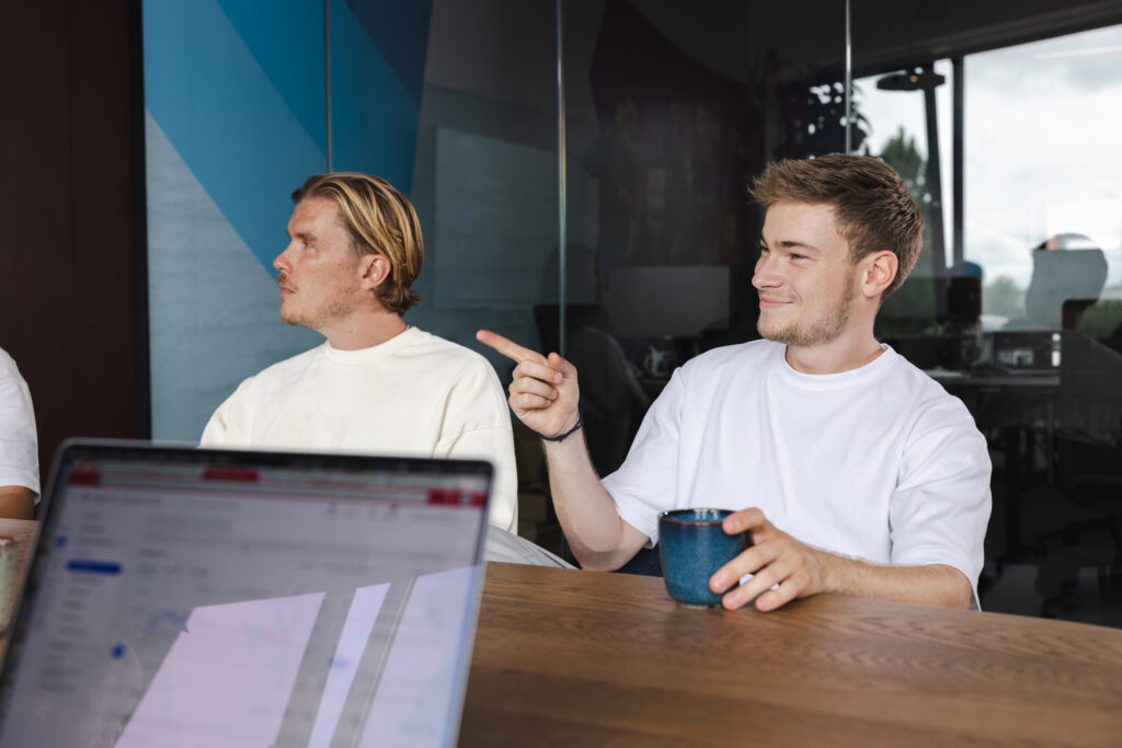 2 collega's die aan een vergadertafel zitten met een koffie in de hand en wijzend naar het scherm